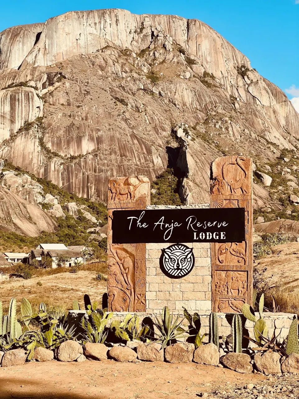 Entrée de The Anja Reserve Lodge à Ambalavao, avec le logo officiel RAGON représentant une tête de lémur catta, face aux montagnes de la Réserve d’Anja sur la RN7 à Madagascar.